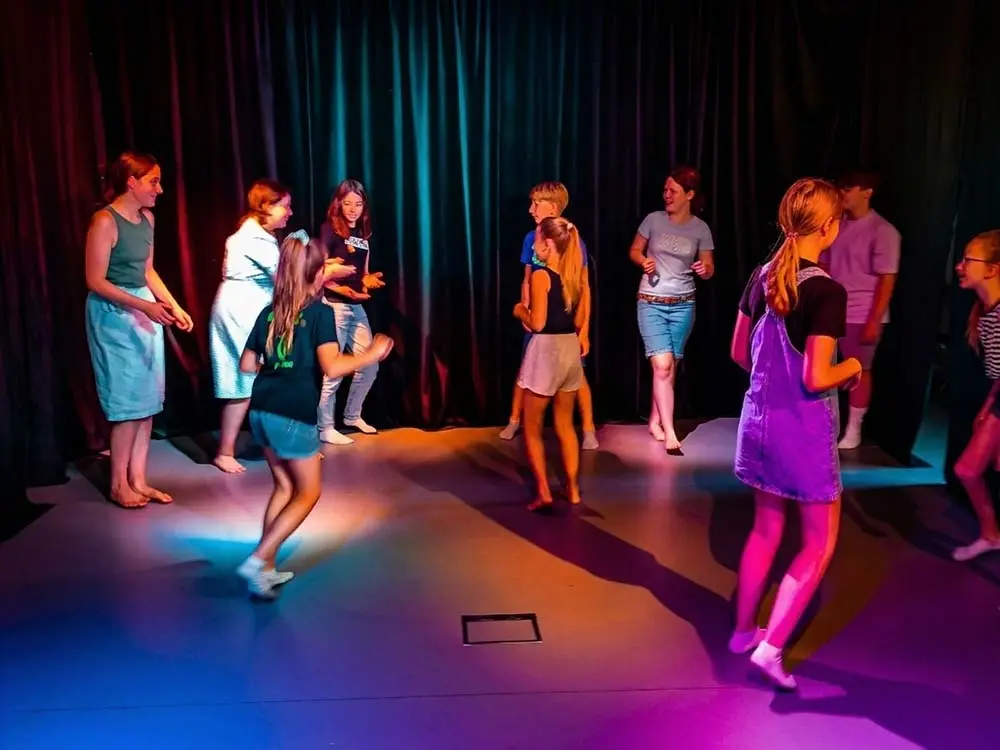 Gruppe von Jugendlichen übt auf einer Theaterbühne; sie stehen im Kreis und bewegen sich frei unter buntem Bühnenlicht in Pink und Blau