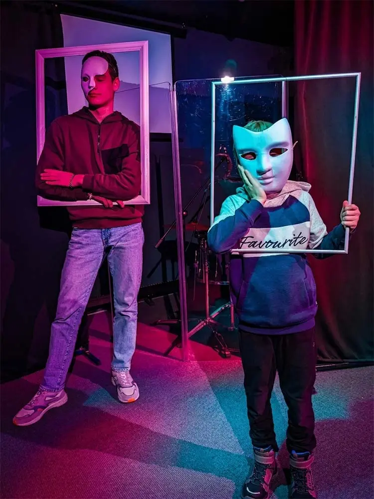 Zwei Jugendliche in einer Theaterszene tragen Theatermasken und halten weiße Bilderrahmen