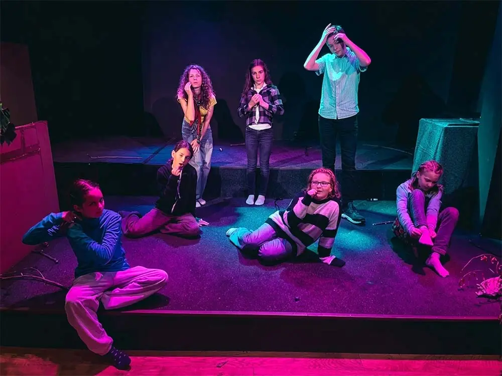 Sieben Kinder und Jugendliche auf einer Theaterbühne in dramatischen Körperposen unter violettem Bühnenlicht; einige sitzen, andere stehen